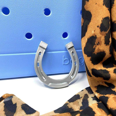 A silver horseshoe stands upright on a blue surface beside animal-print fabric—perfect inspiration for BOGLETS - RODEO Charms by Chatelet Manufacturing, decorative tote bag charms made in the USA to accessorize your favorite Bogg Bag.