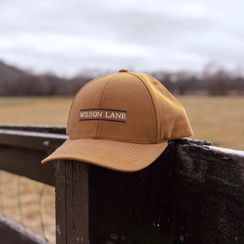 A Down the Lane Structured Dad Hat by Wilson Lane Apparel, featuring a tan chino twill and branded patch, rests on a dark wooden fence with an open field and cloudy sky in the background.