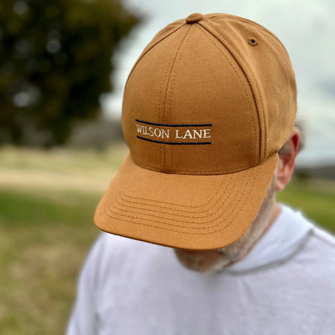 A person in a tan Down the Lane Structured Dad Hat by Wilson Lane Apparel, with the logo embroidered, looks down outdoors in a grassy area. Their face is obscured and they wear a light gray hoodie; the background is blurred.