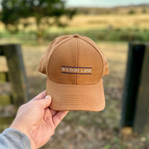 A hand holds the Down the Lane Structured Dad Hat by Wilson Lane Apparel, featuring a rectangular WILSON LANE patch, against a blurred outdoor background of pasture, trees, and fencing.