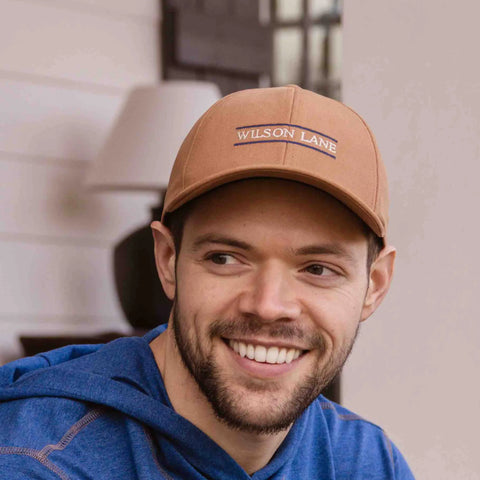 A bearded man smiles indoors wearing a brown Down the Lane Structured Dad Hat by Wilson Lane Apparel and a blue hoodie, with a blurred lamp and wall in the background.