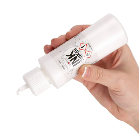 A hand squeezes a white plastic bottle labeled Alcohol Ink Sinker from ArtResin, dispensing a drop of White Alcohol Ink Sinker for making petri art coasters. The label features hazard symbols.