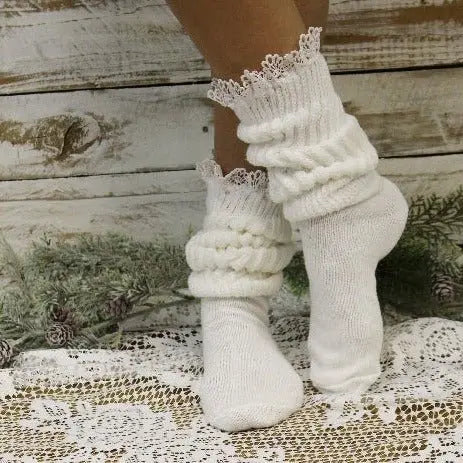 Someone wears Catherine Cole’s LACE Topped Ultimate Slouch Socks in white, standing on a lace-covered surface near rustic wooden boards and greenery.