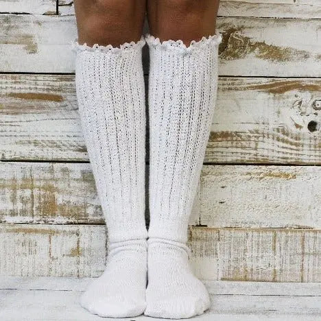 Someone wears Catherine Cole LACE Topped Ultimate Slouch Socks in white, standing on a wooden floor against a rustic white wooden wall.