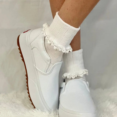 A person wears Catherine Coles HEART Lace Cuff Socks in white, paired with white slip-on platform sneakers, standing on a white fuzzy surface against a neutral background.