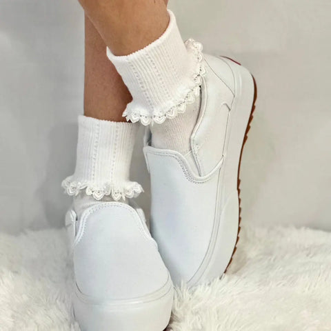 A person wearing Catherine Cole HEART Lace Cuff Socks - White and white slip-on sneakers stands on a fluffy white surface, feet crossed at the ankles.