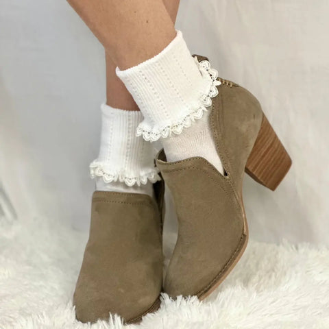 A person wears Catherine Cole HEART Lace Cuff Socks in white with tan suede heeled ankle boots, standing on a soft white fuzzy surface against a light background.