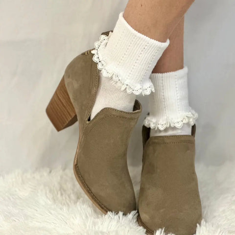Someone wearing Catherine Cole’s HEART Lace Cuff Socks in white pairs them with beige suede heeled ankle boots, standing on a soft white furry rug against a plain background.
