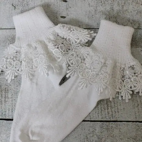 The Catherine Cole SIGNATURE Lace Cuff Socks - White feature ribbed cuffs and intricate white ruffle lace details, displayed on a rustic white wooden surface.
