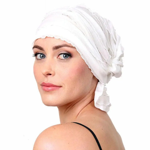 A woman with light skin and blue eyes models the Chemo Beanies® Stella, a white slip-on chemo headscarf by Chemo Beanies®, paired with a black spaghetti-strap top, looking over her shoulder against a white background.