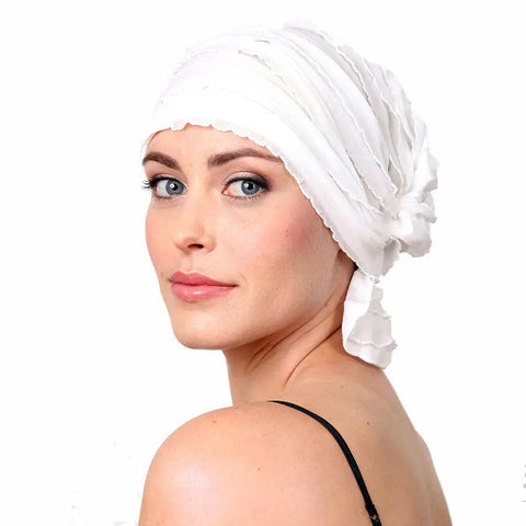 A woman with blue eyes and light skin wears the STELLA CHEMOBEANIES® by Chemo Beanies®, styled as a white headscarf for hair loss, paired with a black spaghetti strap top against a plain white background.