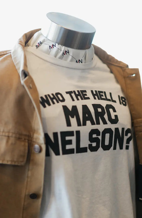 A mannequin sports Marc Nelson’s Who the Hell? Tee—a white, premium cotton shirt with bold black text—layered under a tan jacket and finished with a white collar featuring MN lettering.
