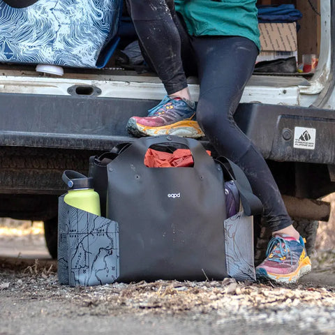 A person in colorful running shoes and black leggings stands by an open van. Nearby is a black eqpd WideTote - Methow Map bag with organized compartments for outdoor gear, a water bottle, and a rolled jacket—ideal for road trips.