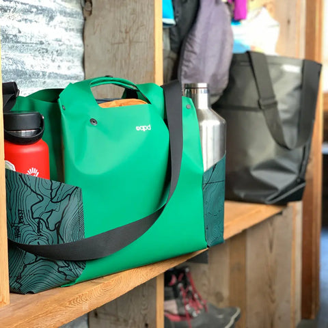 The eqpd WideTote - Methow Map, a bright green bag with black straps, organizes silver and red bottles on a wooden shelf beside a black tote—ideal for road trips in rustic settings with coats hanging above.