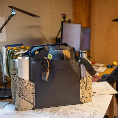The eqpd WideTote - Methow Map, featuring map designs and roomy compartments, holds a water bottle, rolled paper, and glasses on a cluttered desk. Ideal for organizing essentials at work or on the go, while someone works at a computer nearby.