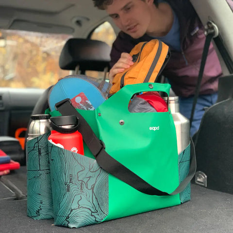 A person packs the eqpd WideTote - Methow Map, a green map-patterned tote with compartments, loading water bottles and gear into the car for road trips and outdoor adventures.
