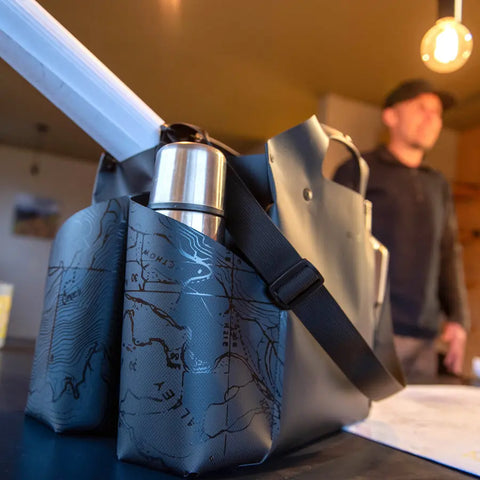 The eqpd WideTote - Methow Map is a grey tote bag featuring a map design and compartments to organize essentials like a thermos and documents, shown in close-up with a casually dressed man blurred in the warmly lit background.