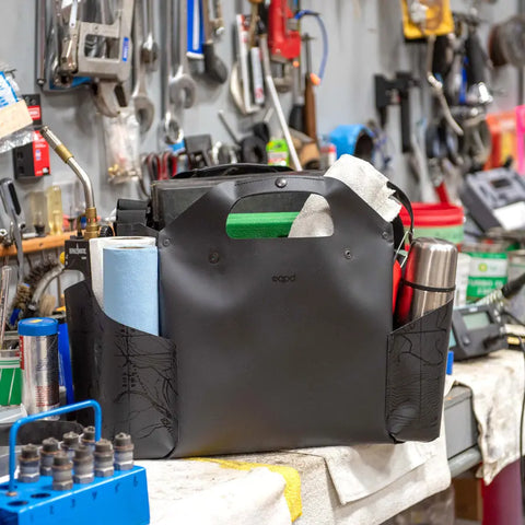 The eqpd WideTote - Methow Map is a black organizer with compartments and side pockets that holds paper towels, a thermos, and more, helping keep your workbench neat in a tool-packed workshop.