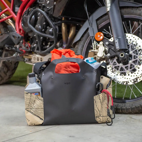 The eqpd WideTote - Methow Map is a black and tan gear bag with a topographic map design, shown beside a motorcycle, featuring multiple compartments to organize essentials—ideal for road trips.