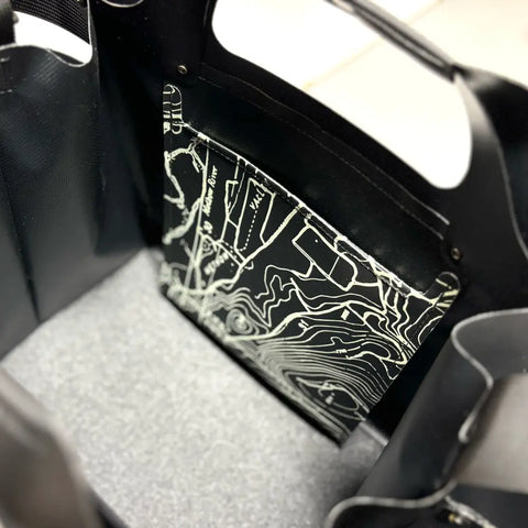 A close-up of the eqpd WideTote - Methow Map shows compartments and a pocket with a black-and-white contour map lining—ideal for organized road trips. The black bag is placed on a gray textured surface.