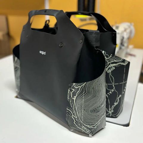 The eqpd WideTote - Methow Map is a black tote bag with short handles and side pockets featuring white map lines, shown on a white surface in a workshop—ideal for organizing essentials on road trips.