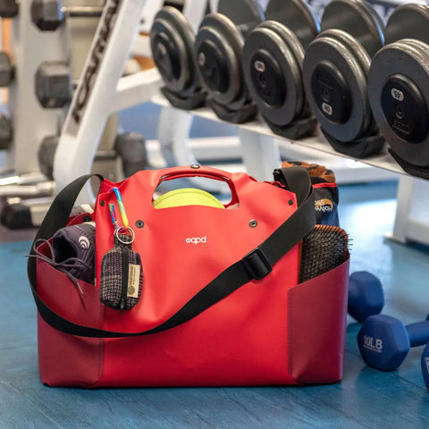 The eqpd WideTote gym bag in red sits on a blue floor with dumbbells and gear nearby. A yoga mat, towel, and water bottle are visible—ideal for workouts or road trips thanks to its unique compartments.
