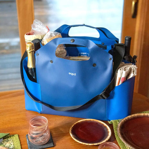 An eqpd WideTote in blue, filled with groceries like a baguette, wine, and wrapped food, sits on a wooden table set with glasses, plates, and green napkins by a window.