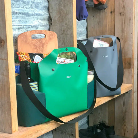 Two reusable eqpd WideTote bags, one green and one gray, are filled with groceries on a wooden shelf. Their convenient compartments make these shopping bags ideal for organizing items in rustic kitchens or packing for road trips.