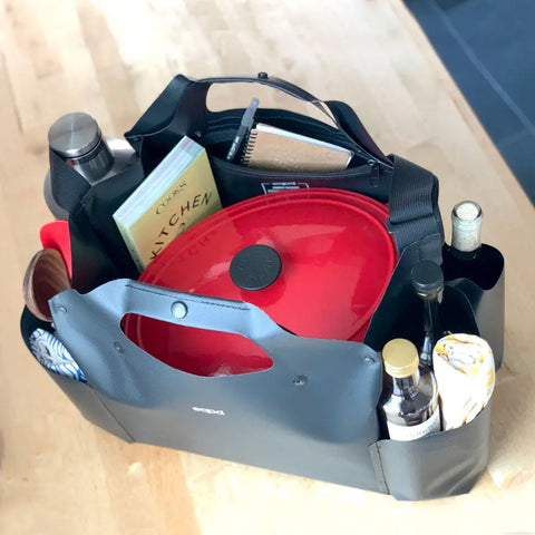 The eqpd WideTote, shown in black and packed with essentials like a red-lidded pot, cookbook, water bottle, oil or vinegar bottles, notebook, and cloth on a wooden surface, is perfect for organized road trips with its unique compartments.