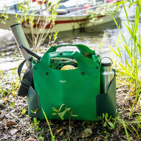 The eqpd WideTote outdoor bag in green sits on grass by a lake, holding gear and a bottle in its side pockets. Surrounded by leafy plants, its ideal for road trips, with a boat and water visible in the background.