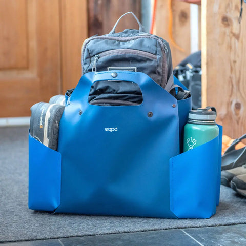 A blue eqpd WideTote with unique compartments neatly holds shoes, a water bottle, and a gray backpack on the floor by a wooden door and post—ideal for organized road trips.