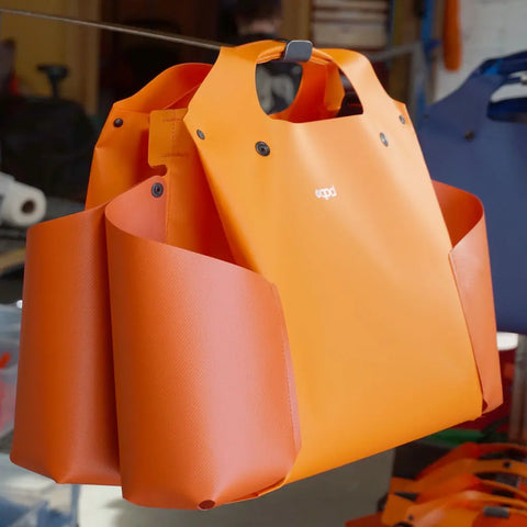 A vibrant orange eqpd WideTote with unique compartments and wide side pockets hangs in a workshop, featuring the white eqpd logo on the front. Ideal for road trips, a blue WideTote is blurred in the background.