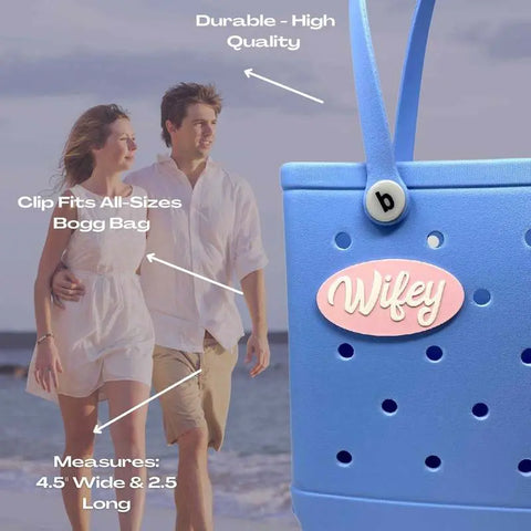 A couple walks on the beach in light clothing. In the foreground, a Chatelet Manufacturing FRESHe BOGLETS Wifey Charm (pink and white) decorates a blue tote—high quality, universal clip, fits most totes, measures 4.5 x 2.5.