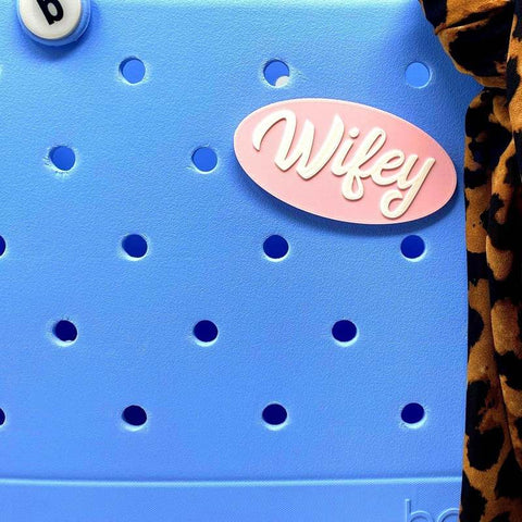 A pink and white Wifey charm by Chatelet Manufacturing, compatible with Bogg Bags, Simply Southern bags, and other tote bags; shown on a light blue perforated tote with leopard print fabric draped over the right edge.