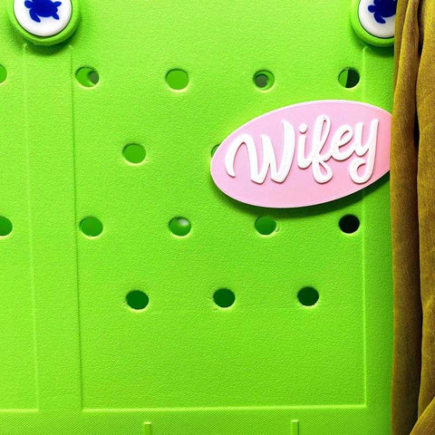 A bright green perforated board displays a Chatelet Manufacturing FRESHe BOGLETS Wifey Charm (pink and white), compatible with Simply Southern, Bogg, and other tote bags. Two blue clover buttons and brown fabric hang on the right side.