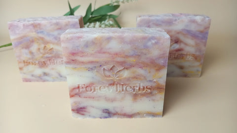 Three rectangular bars of Wild Orchid Handmade Soap by Forev'Herbs in marbled white, pink, yellow, and purple tones, each embossed with the brand name and leaf logo. Blurred green leaves and white flowers appear in the background.