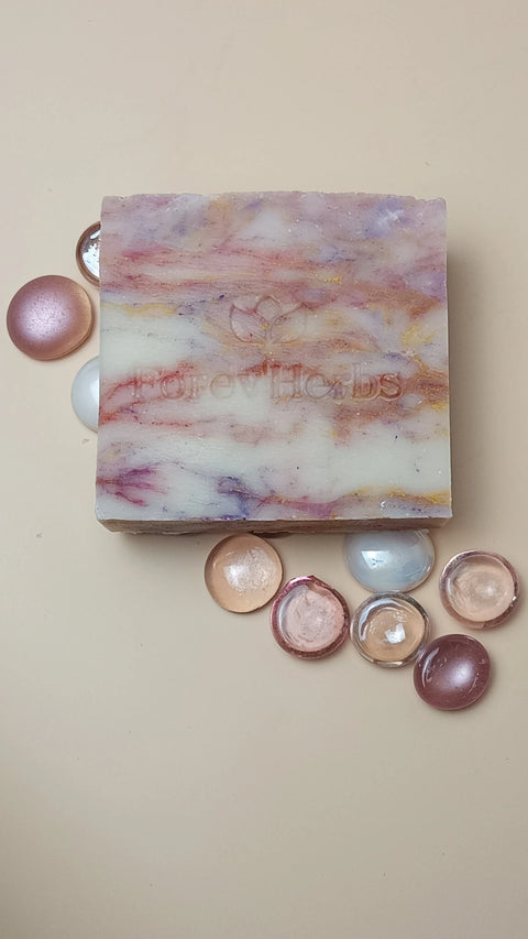 A marbled Wild Orchid Handmade Soap by Forev'Herbs, with pink, yellow, and purple swirls, rests among pastel and metallic pebbles on a beige surface—bringing moisturizing luxury to your bath decor.