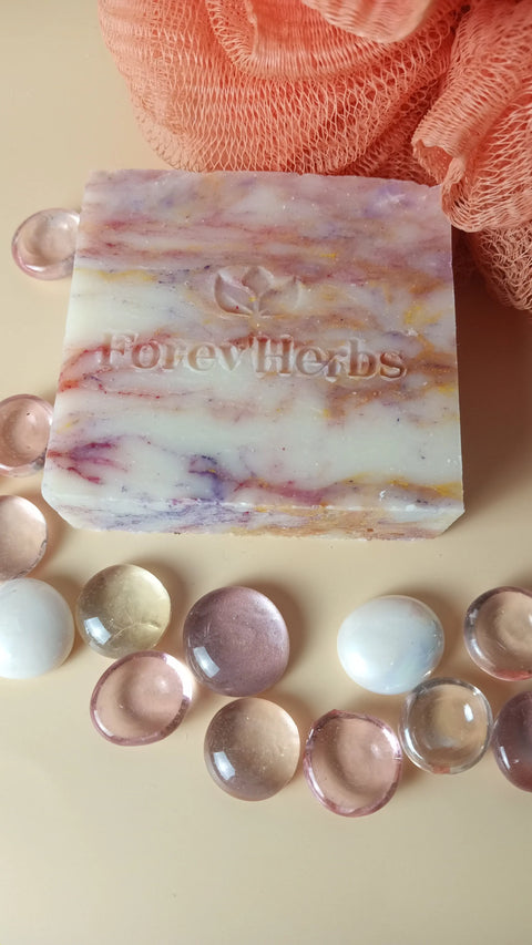 A marbled Wild Orchid Handmade Soap bar by Forev'Herbs, engraved with the brand name, sits among clear and colored glass pebbles beside an orange bath sponge on a beige surface.