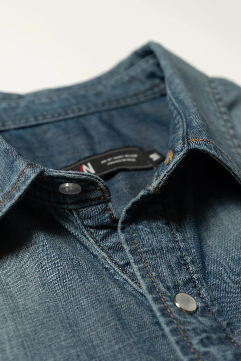 Close-up of a blue Marc Nelson Wild West Pearl Snap Shirt, highlighting the collar, metal snap buttons, visible stitching, and a partially visible black tag inside the collar—showcasing this classic Western style.