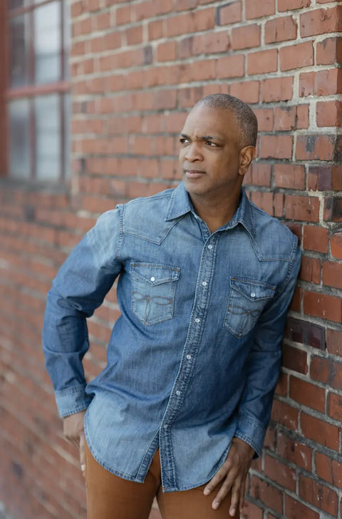 A man in the Marc Nelson Wild West Denim Pearl Snap Shirt—rugged Western style, made in the USA—and brown pants leans against a brick wall, gazing thoughtfully. A red brick building with a window is visible behind him.