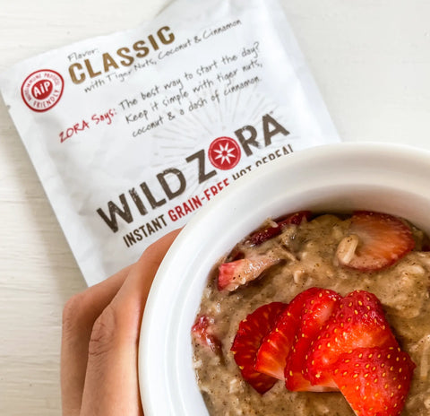 A hand holds a bowl of hot cereal topped with fresh strawberries. Behind it is a Wild Zora Instant Cereal - AIP Classic With Tiger Nuts, Coconut & Cinnamon 5-Pack, made for the autoimmune protocol diet.