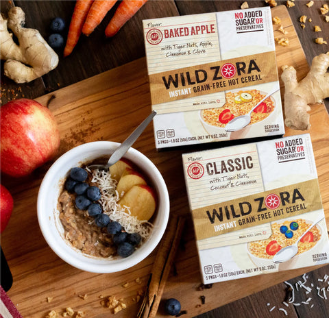 A hot bowl of Wild Zora Instant Cereal - AIP Multi-Pack Classic & Baked Apple 10-Pack, topped with blueberries, apple slices, and shredded coconut sits next to two boxes, apples, carrots, ginger, and a striped cloth on a wooden table.