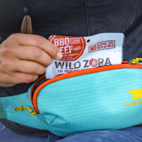 Someone holds a pack of Wild Zora Bars - BBQ Beef With Kale, Tomato & Red Bell Pepper 10-Pack, partially pulled from a blue zippered pouch. The packaging notes 100% grass-fed beef and no gluten, soy, nuts, or milk.