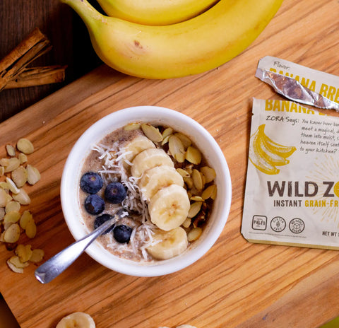 A bowl of oatmeal topped with banana, blueberries, coconut, almonds, and flaxseed sits beside Wild Zora Instant Cereal - Banana Bread With Bananas, Almonds & Cacao 10-Pack, a spoon, a banana, cinnamon sticks, and extra almond slices on a wooden board.