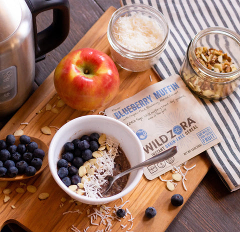 A bowl of Wild Zora Instant Cereal - Blueberry Muffin With Blueberry, Almond & Coconut sits on a wooden board, topped with blueberries, coconut, and almonds. An apple, loose blueberries, nuts, and a Wild Zora 10-Pack packet are nearby.