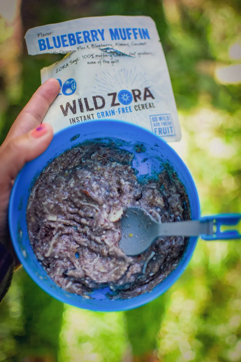A hand holds a blue bowl of Wild Zora Instant Cereal - Blueberry Muffin With Blueberry, Almond & Coconut, with a spoon and open 10-pack packaging above it, set against blurred greenery. Made with organic flaxseed.