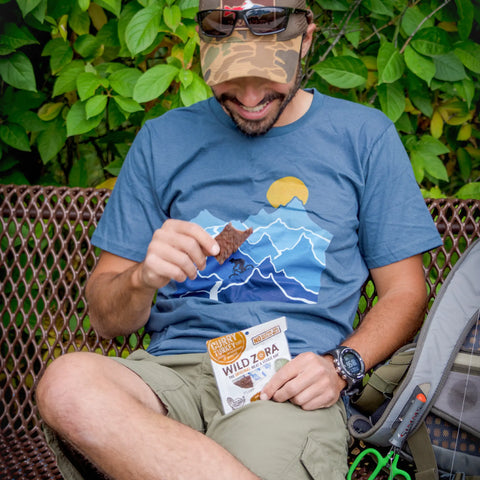 A man in a blue mountain-print shirt and camo hat sits on a bench outdoors, smiling as he holds a Wild Zora Bars - Curry Turkey With Spinach, Dates & Cardamom 10-Pack. A backpack and greenery nearby reflect his love for Wild Zora meat & veggie bars.