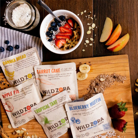 A bowl of yogurt with berries and granola sits beside sliced apples, scattered fruit, and five Wild Zora Instant Cereal - Variety 5-Pack packets—perfect for a high fiber breakfast—on a wooden surface.