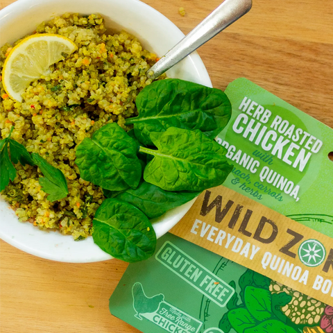 A bowl of Wild Zora Quinoa Meals – Herb Roasted Chicken With Spinach, Carrots & Herbs sits beside its packaging on a wooden surface—a quick, wholesome quinoa meal ready for any time.