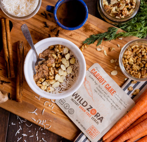 A bowl of Wild Zora Instant Cereal - Carrot Cake With Carrots, Raisin & Clove sits on a wooden board with nuts, coconut, almonds, a spoon, carrots, coffee mug, cinnamon sticks, grated coconut, and its 10-pack cereal packet.